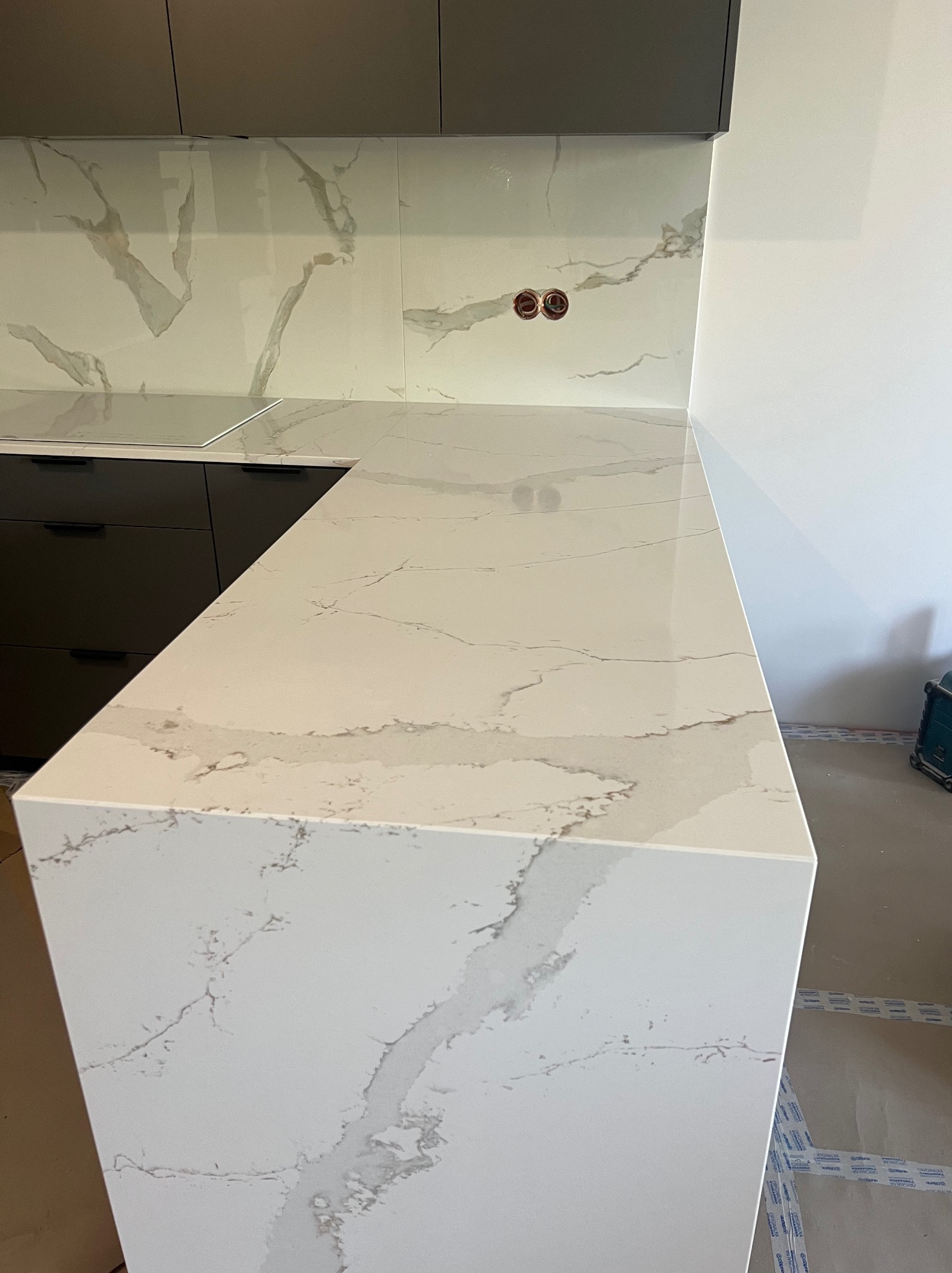 Biały blat kuchenny z marmurowym wzorem, dopasowany do płytek na ścianie, widok na czarne szafki i gniazdka elektryczne, ukazujący estetyczne wykończenie.