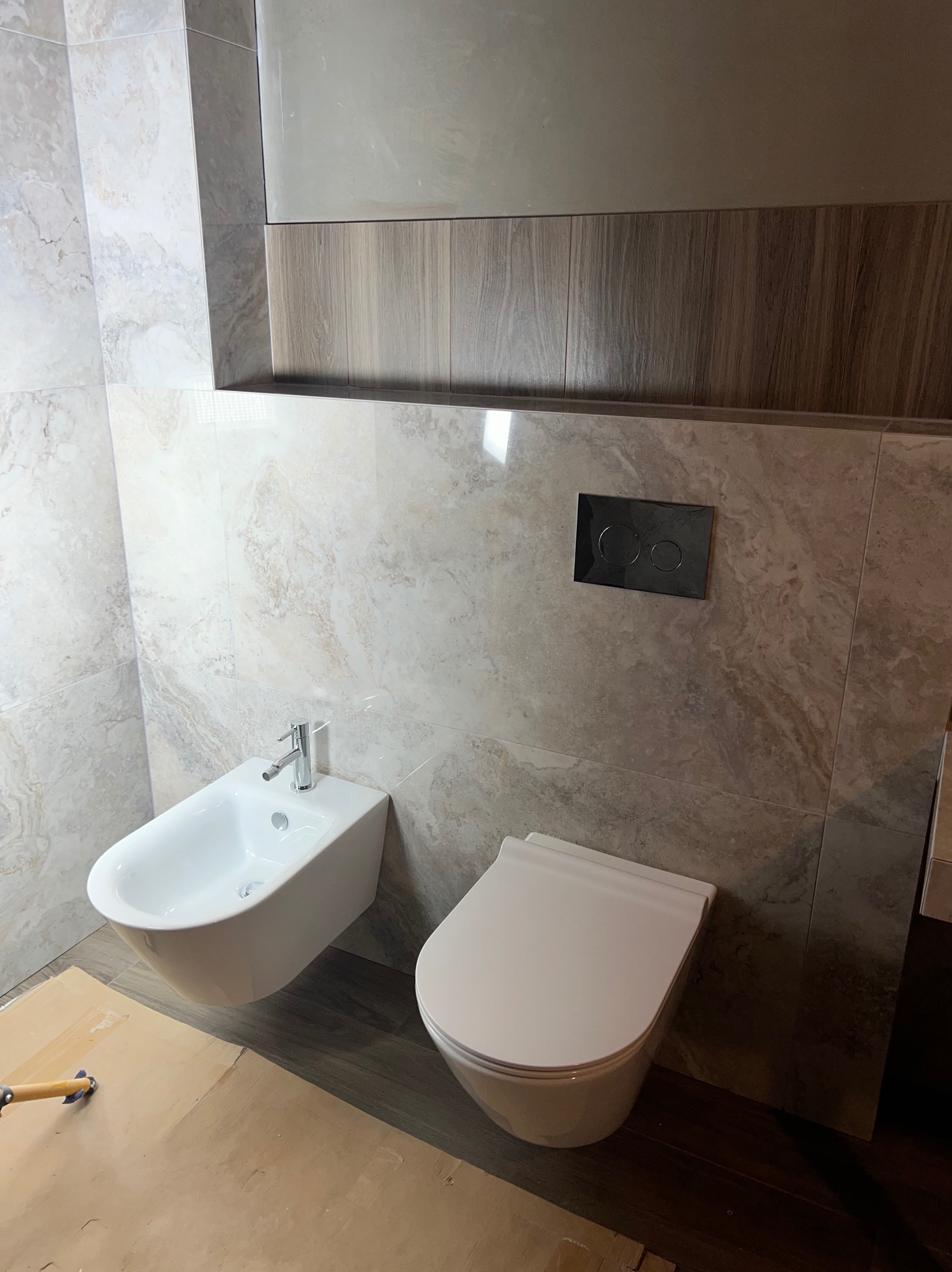 Wykończona łazienka z marmurowymi płytkami, wiszącą toaletą i bidetem. Widoczny fragment podłogi zabezpieczony kartonem. Nowoczesny design z akcentami drewna.