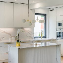 HODER - Jasna kuchnia z marmurowym blatem i wyspą. Złote akcenty: bateria, uchwyty, lampa. Białe szafki i sprzęt AGD. Minimalistyczny design, kwiat w ozdobnej doniczce.