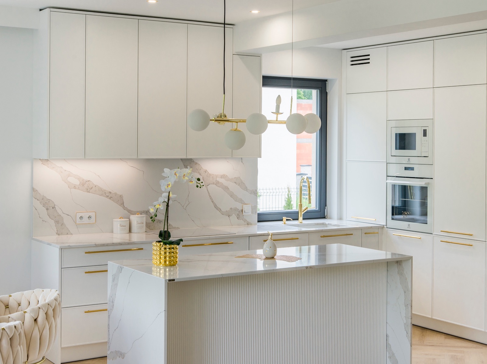 Jasna kuchnia z marmurowym blatem i wyspą. Złote akcenty: bateria, uchwyty, lampa. Białe szafki i sprzęt AGD. Minimalistyczny design, kwiat w ozdobnej doniczce.
