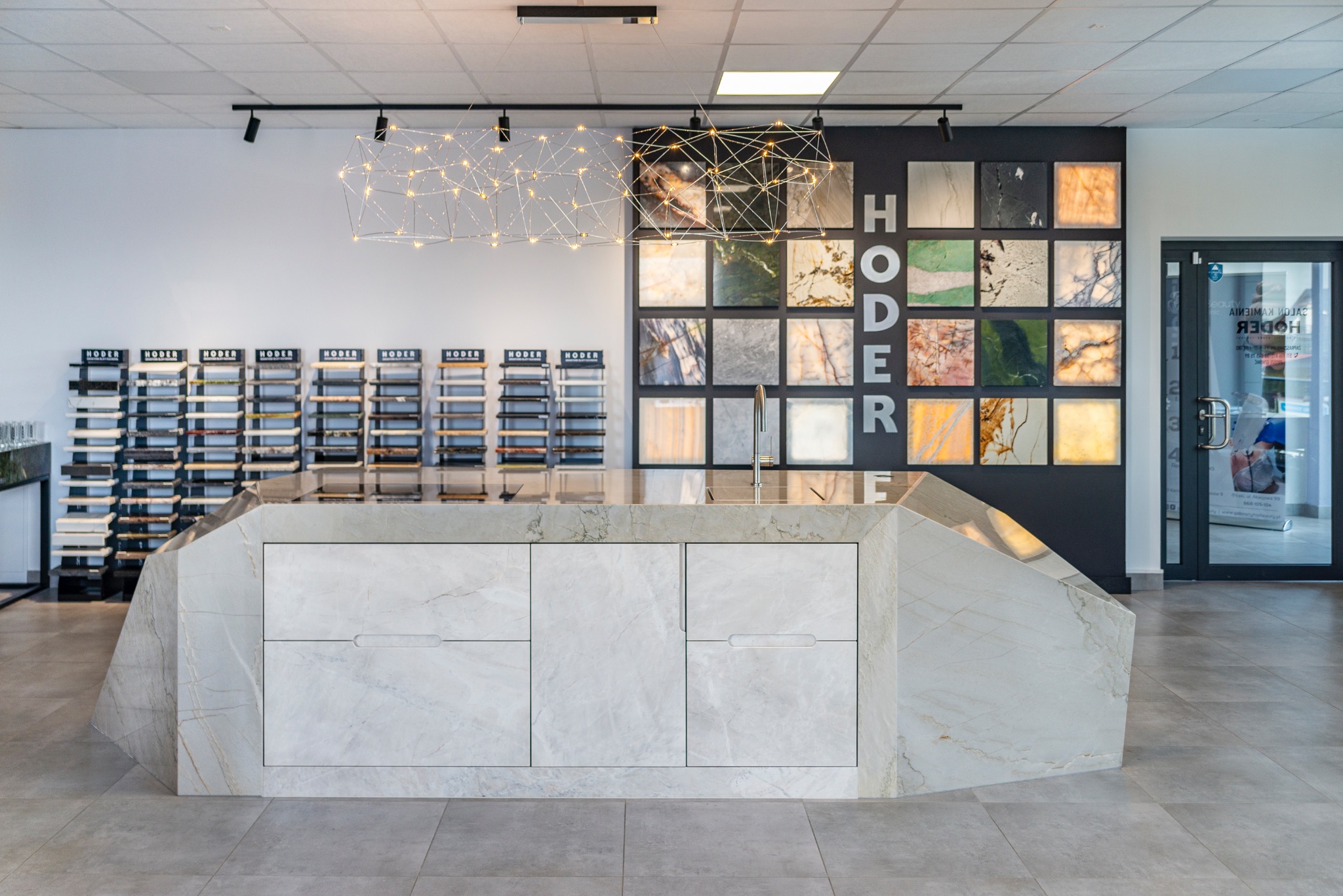 Salon z ekspozycją blatów kamiennych: marmurowy blat na wyspie kuchennej, próbki kamieni na ścianie, nowoczesna lampa. Jasne wnętrze, szare płytki podłogowe.