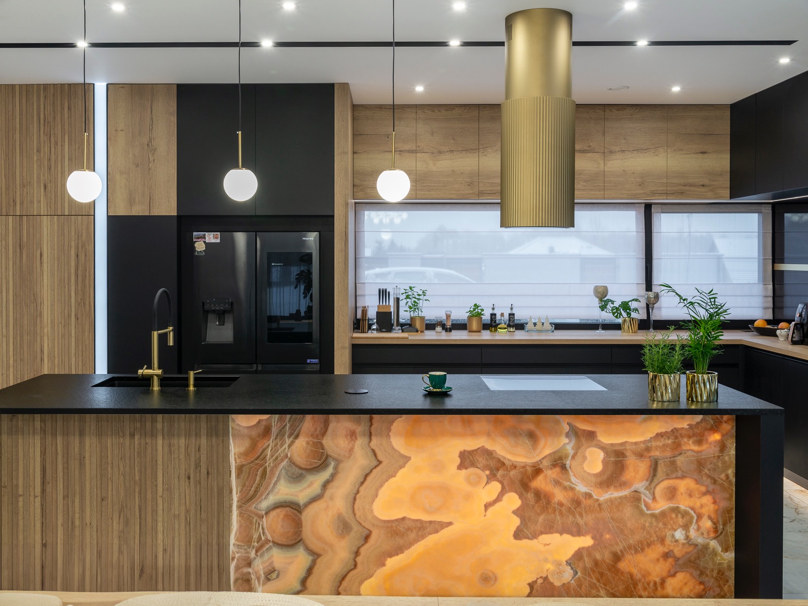 Nowoczesna kuchnia z czarnym blatem i wyspą z podświetlanego kamienia onyksowego. Złote akcenty: armatura, okap i doniczki. Drewniane i czarne szafki.