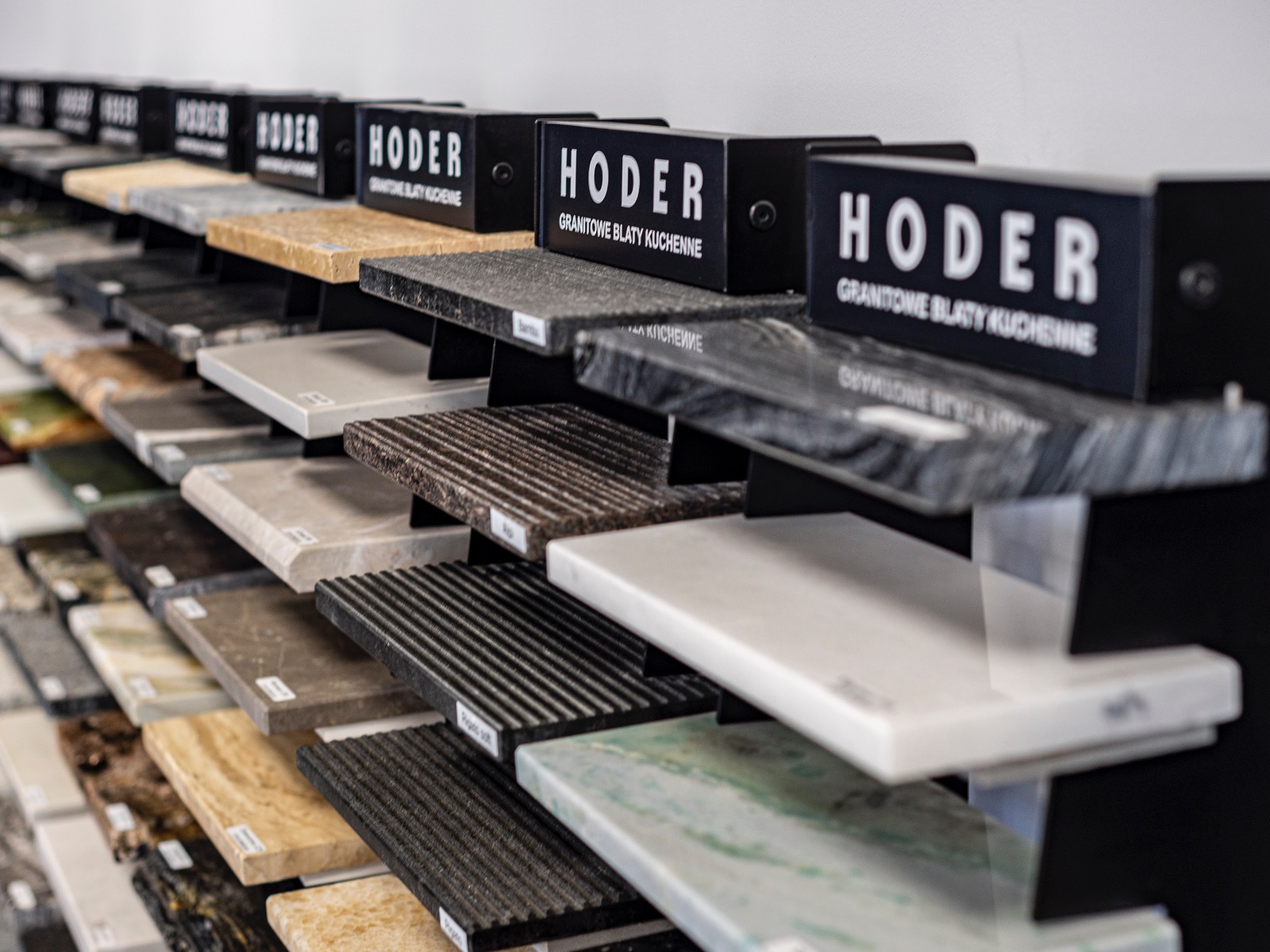 Ekspozycja blatów kamiennych Hoder: różnorodność wzorów i kolorów, od beżu po grafit, z etykietami. Blaty na czarnym regale, oświetlone, widok z perspektywy.