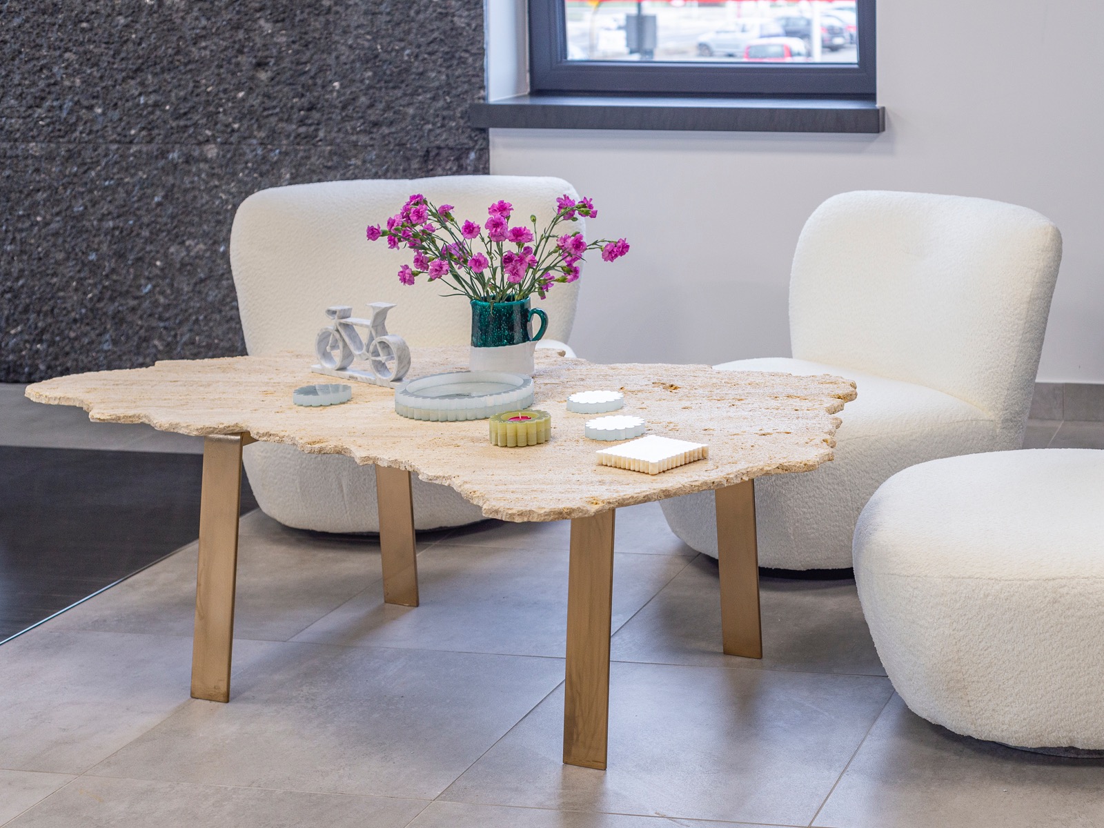 Stolik kawowy z blatem z trawertynu o nieregularnym kształcie i złotymi nogami, otoczony białymi fotelami typu baranek. Na blacie dekoracje i kwiaty.