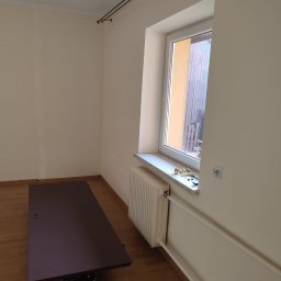 Marek Rząsa - Pokój po malowaniu: jasne ściany, okno z parapetem, grzejnik, karnisz. Na podłodze deski i zabezpieczona powierzchnia. Widoczne narzędzia malarskie.