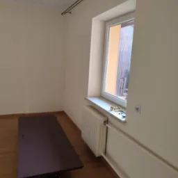 Pokój po malowaniu: jasne ściany, okno z parapetem, grzejnik, karnisz. Na podłodze deski i zabezpieczona powierzchnia. Widoczne narzędzia malarskie.