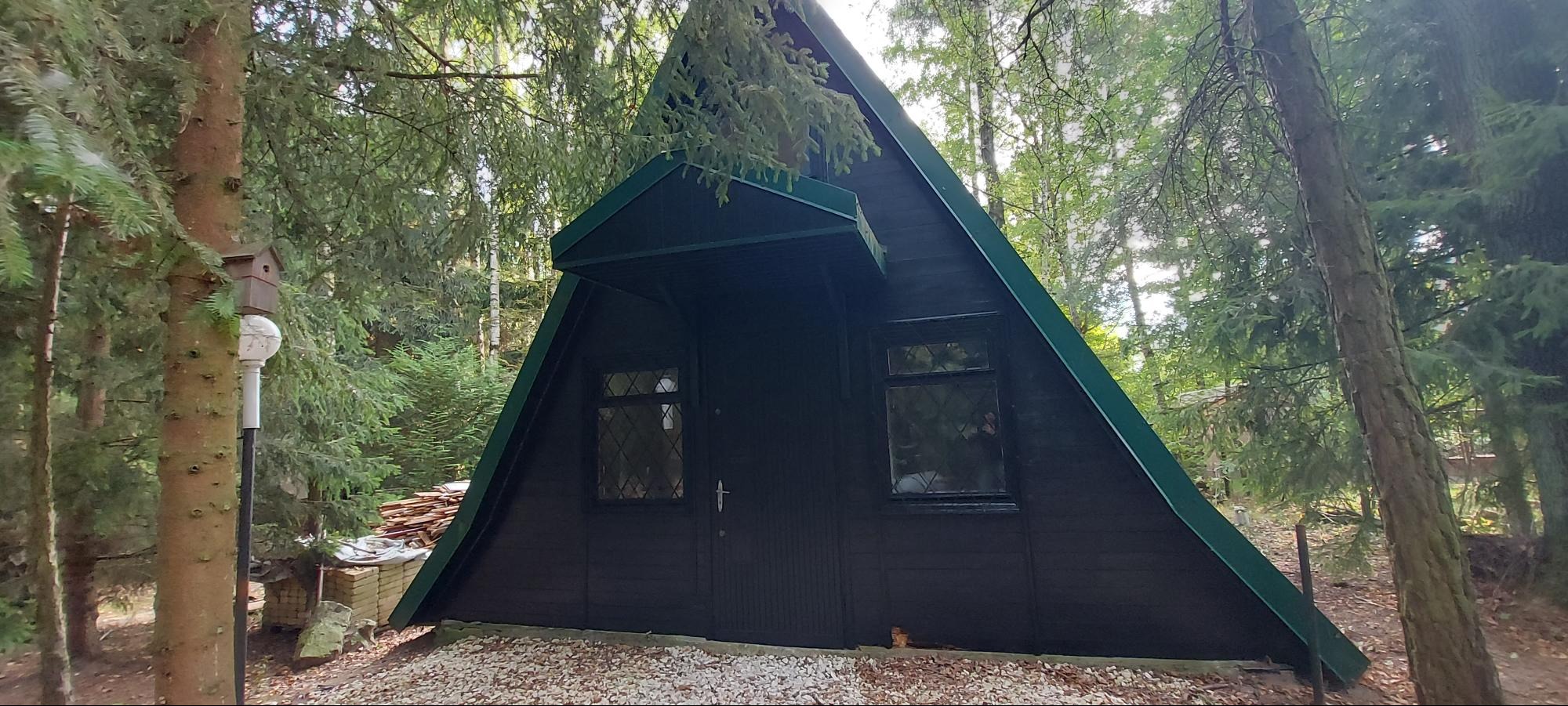 Czarny domek letniskowy w kształcie litery A, z zielonymi akcentami, otoczony drzewami. Widoczna sterta drewna, lampa i budka dla ptaków. Elewacja po renowacji.