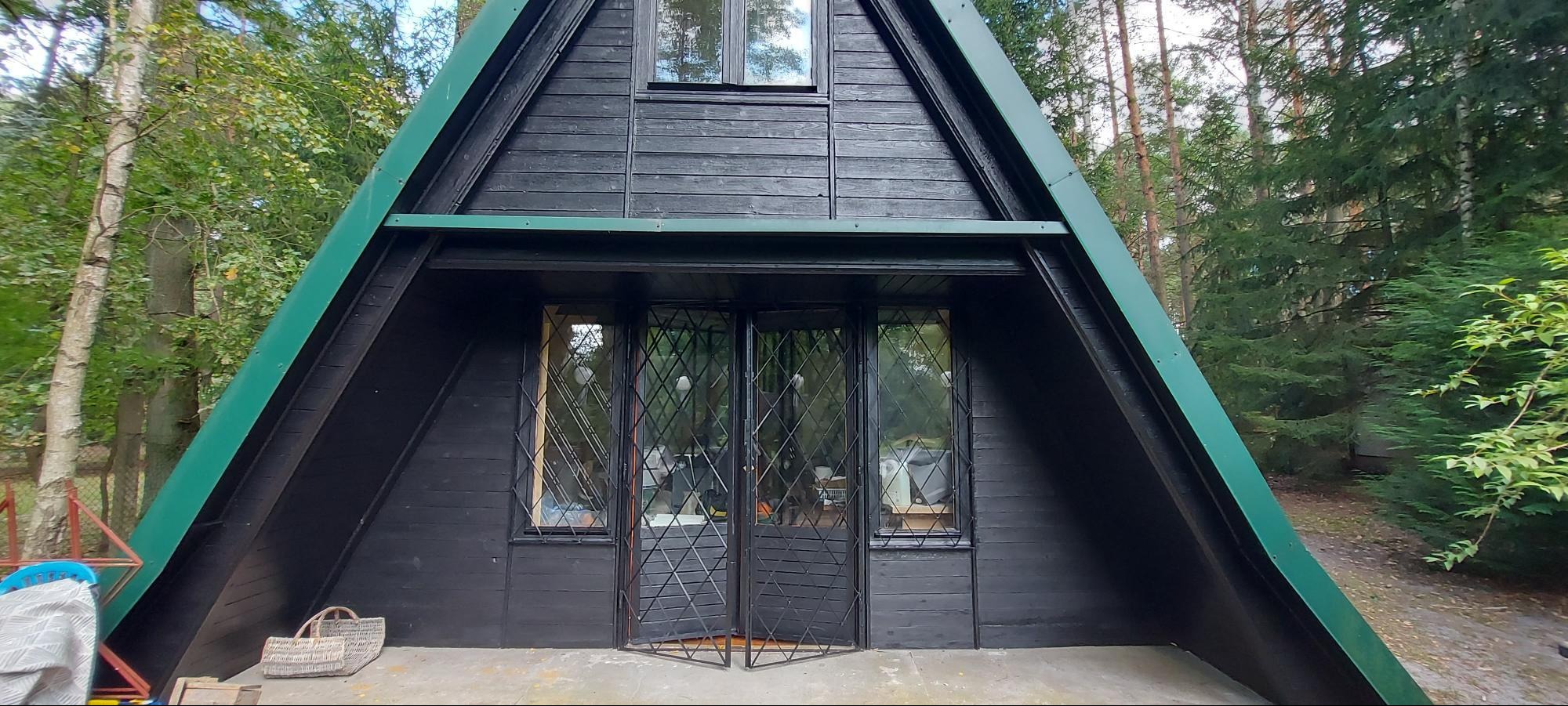 Czarny, drewniany domek letniskowy w kształcie litery A z zielonymi akcentami, widziany z przodu, otoczony zielenią drzew. Widoczne okna i drzwi z kratami.