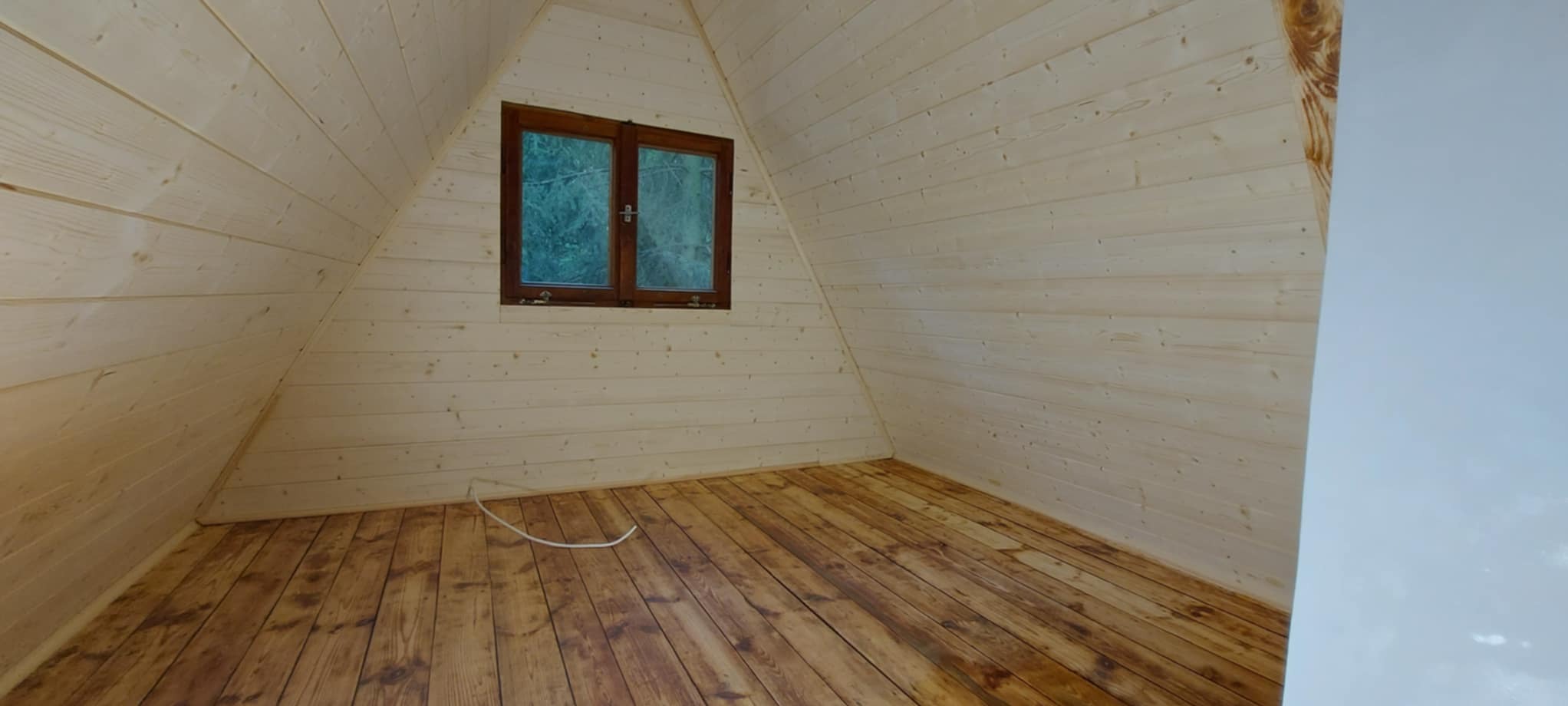 Wnętrze drewnianego domku z oknem i podłogą z desek. Ściany wyłożone jasnym drewnem, widoczny kabel na podłodze.