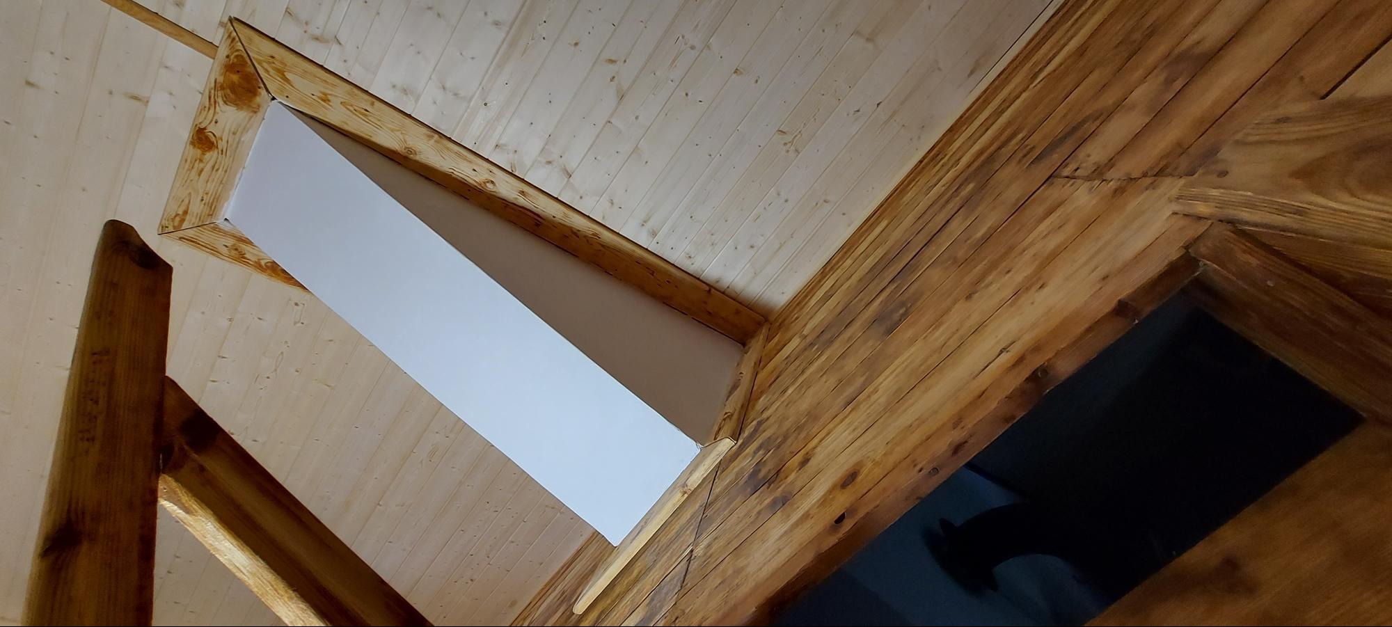 Drewniane wykończenie sufitu z białym, pomalowanym fragmentem. Widok z dołu, ukazujący strukturę drewna i kontrast z bielą. Detale belek i desek.