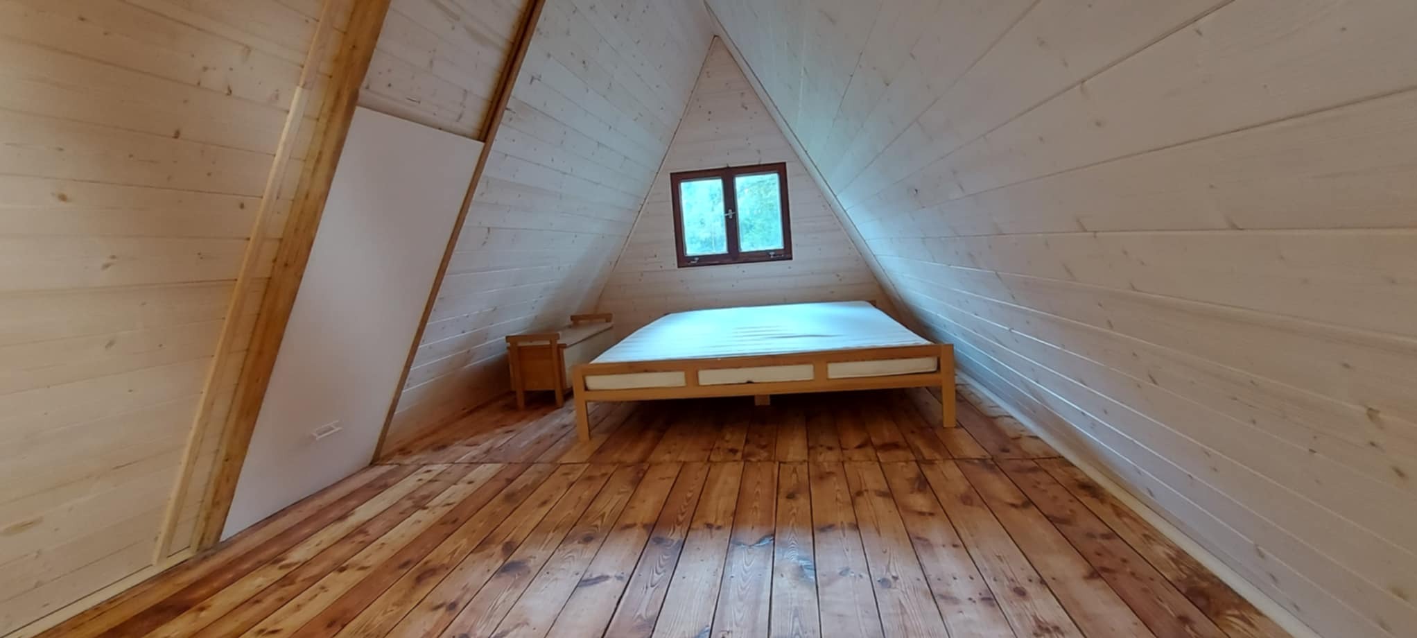 Drewniane wnętrze poddasza z łóżkiem i oknem, ściany i sufit w jasnym odcieniu, podłoga z ciemniejszego drewna. Widok na pomalowane deski.