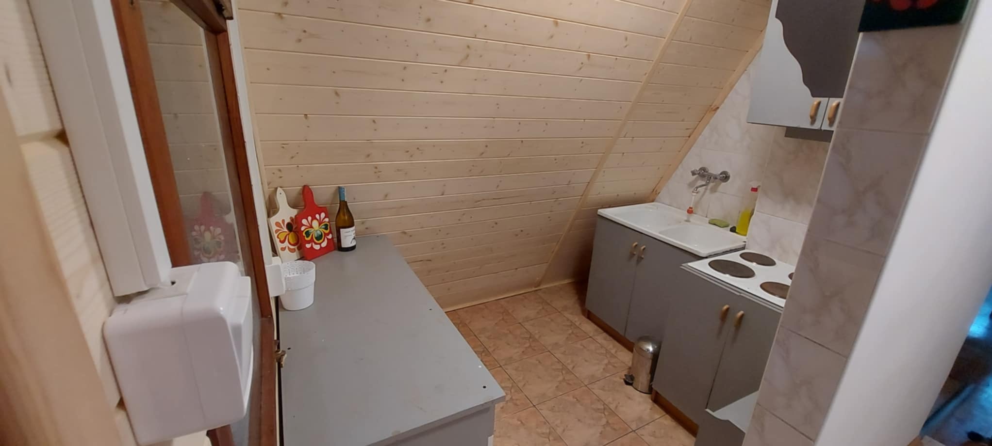 Drewniana kuchnia na poddaszu z szarymi szafkami, białym zlewem i płytą grzewczą. Dekoracyjne deski do krojenia i butelka wina na blacie dodają uroku.