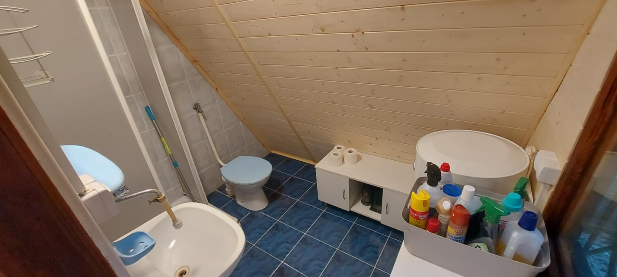Ukończona łazienka na poddaszu z drewnianą boazerią, niebieską glazurą i białą armaturą. Widoczne środki czystości.