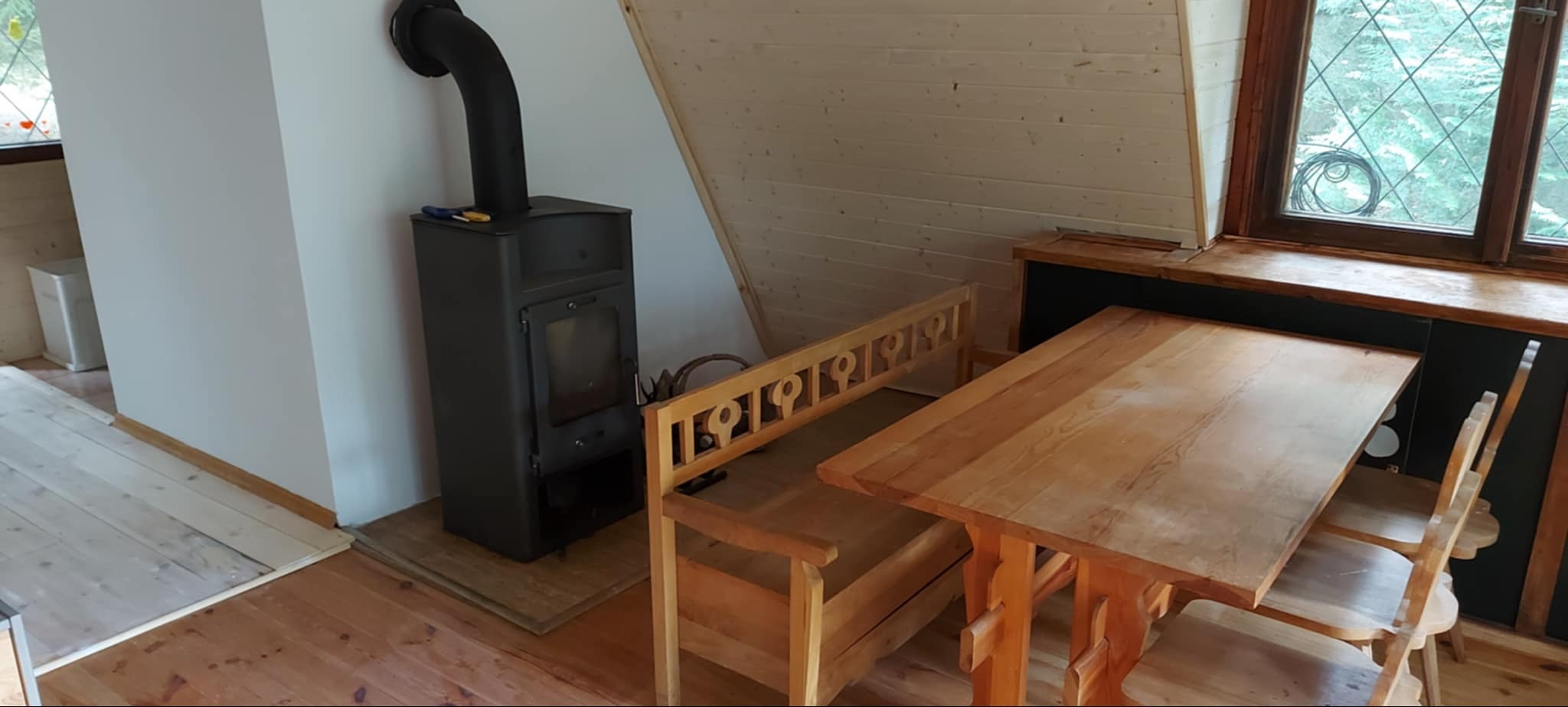 Jasne wnętrze poddasza z drewnianą boazerią, piecem typu koza, drewnianym stołem i ławką. Widoczne świeżo pomalowane ściany.