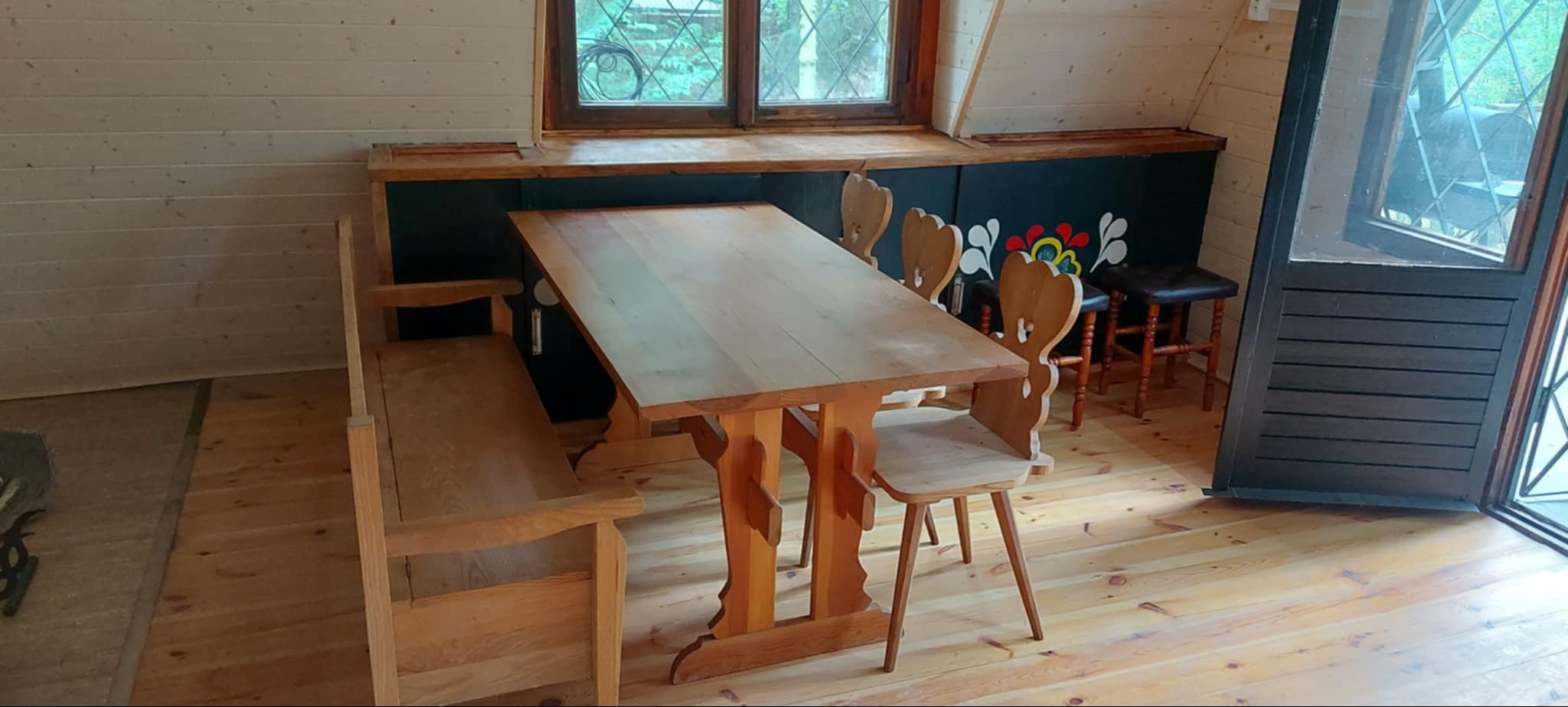 Wnętrze w stylu rustykalnym: drewniany stół z ławą i krzesłami, ściany z boazerii. Dekoracyjne malunki na ścianie pod oknem. Drewniana podłoga. Widok na zieleń za oknem.