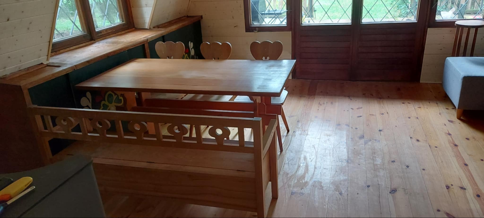 Drewniane wnętrze: stół z ławką i krzesłami w kształcie serc, jasna podłoga, okna z widokiem na zieleń. Ściany z boazerii. Widoczne ślady użytkowania.