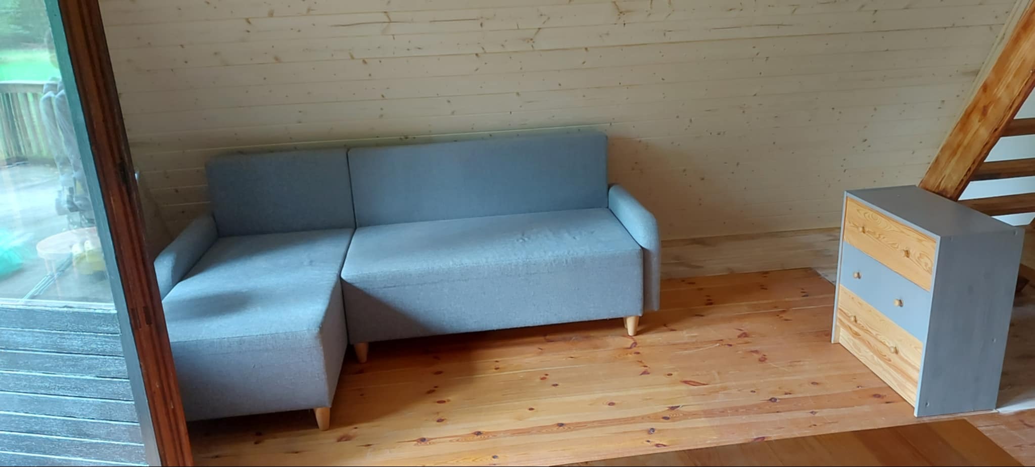 Jasne poddasze: szara sofa narożna i komoda z drewnianymi frontami szuflad. Ściany i sufit wykończone boazerią, podłoga drewniana. Widok na okno i schody.