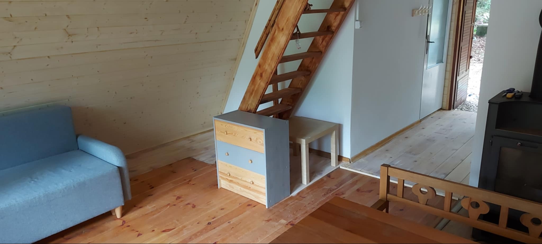 Jasne, drewniane wnętrze domu z niebieską sofą, drewnianymi schodami i komodą w odcieniach szarości. Widoczna podłoga z desek i fragment pieca.