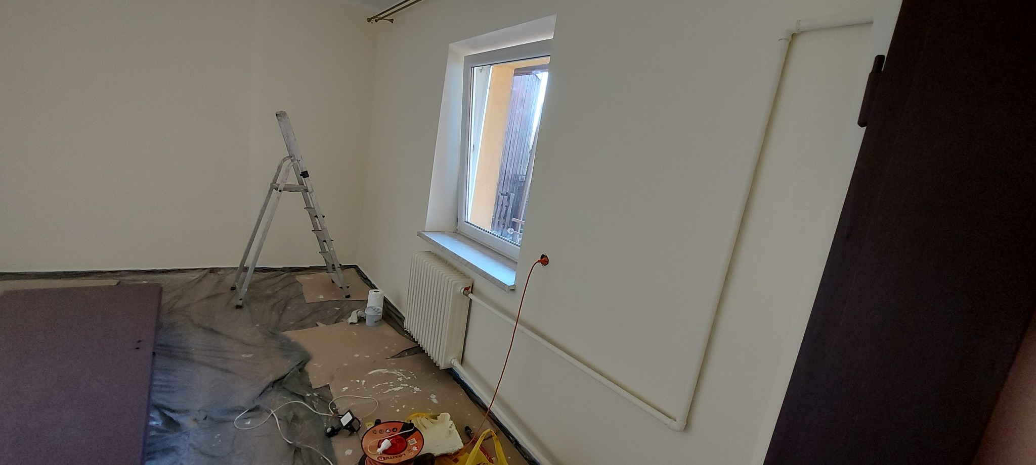 Pokój po malowaniu na biało, z zabezpieczoną podłogą i grzejnikiem. Widoczna drabina malarska i akcesoria malarskie. Świeżo pomalowane ściany.