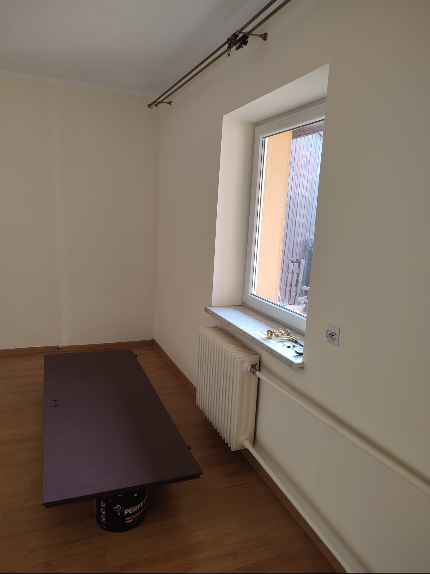 Pokój po malowaniu: jasne ściany, okno z parapetem, grzejnik, karnisz. Na podłodze deski i zabezpieczona powierzchnia. Widoczne narzędzia malarskie.