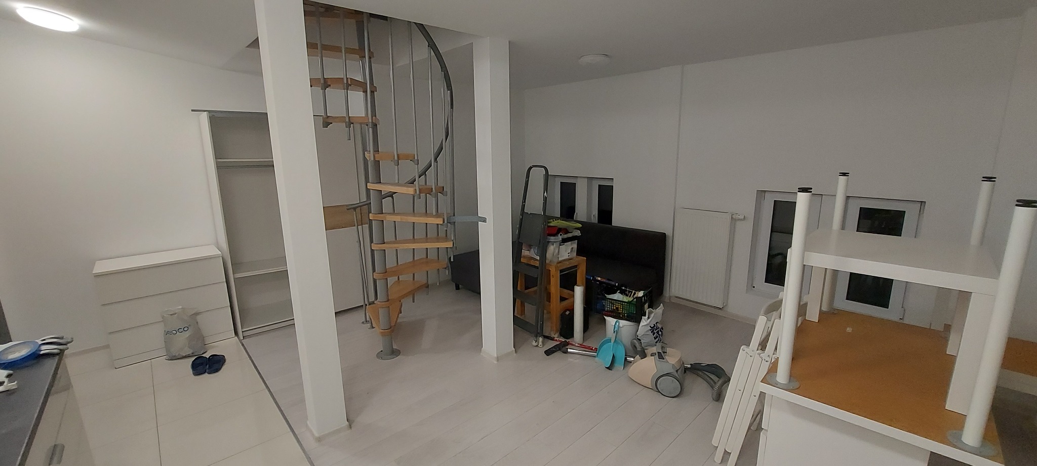 Wnętrze mieszkania przed malowaniem: spiralne schody, białe ściany, meble częściowo odsłonięte, widoczne narzędzia i akcesoria malarskie, przygotowane do prac remontowych.