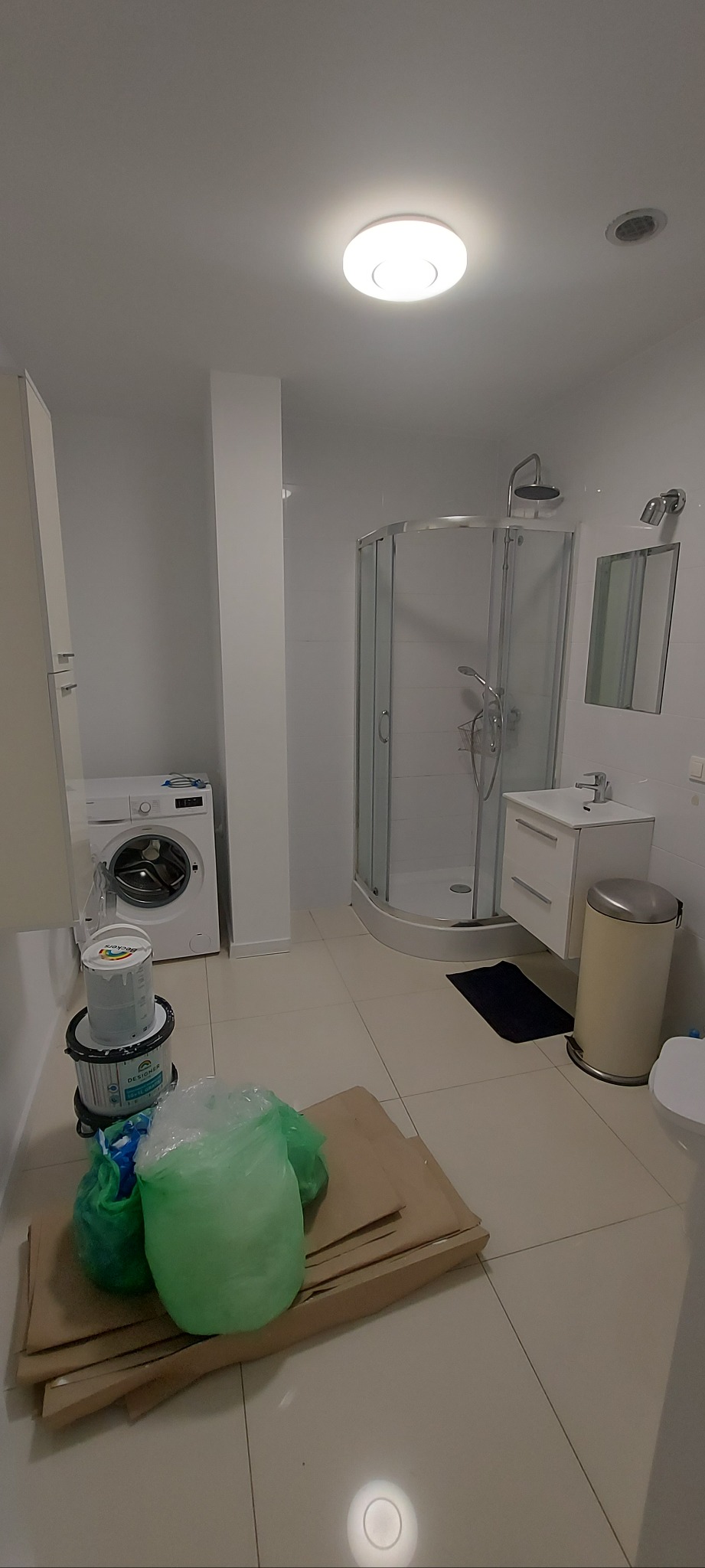 Wnętrze łazienki po remoncie, widoczna kabina prysznicowa, umywalka, toaleta i pralka. Na podłodze stoją farby i zabezpieczenia malarskie.