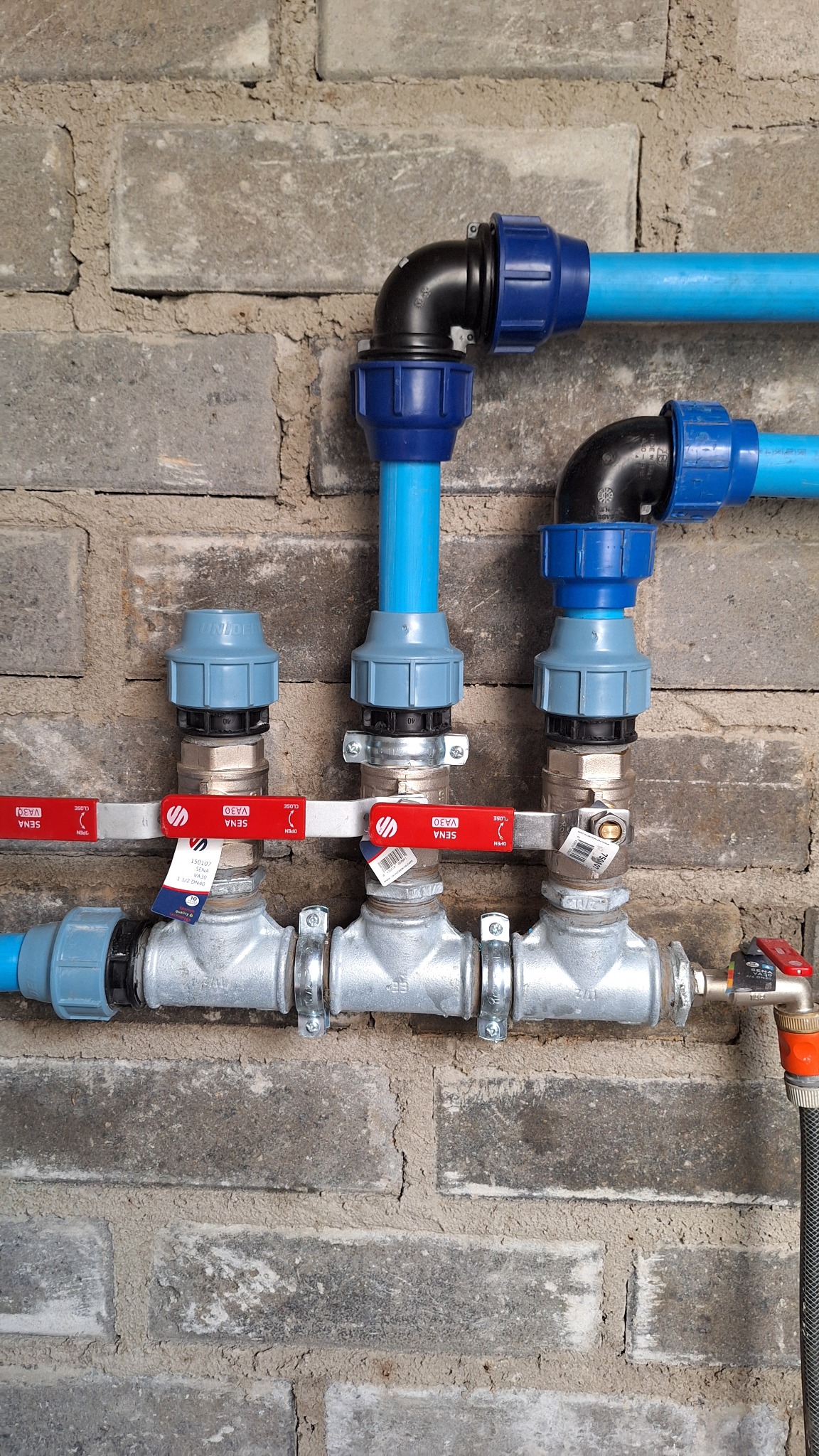 Instalacja wodna z niebieskich rur i srebrnych zaworów marki Sena na tle szarej, ceglanej ściany. Widoczne złączki i etykiety na zaworach. Ujęcie centralne.