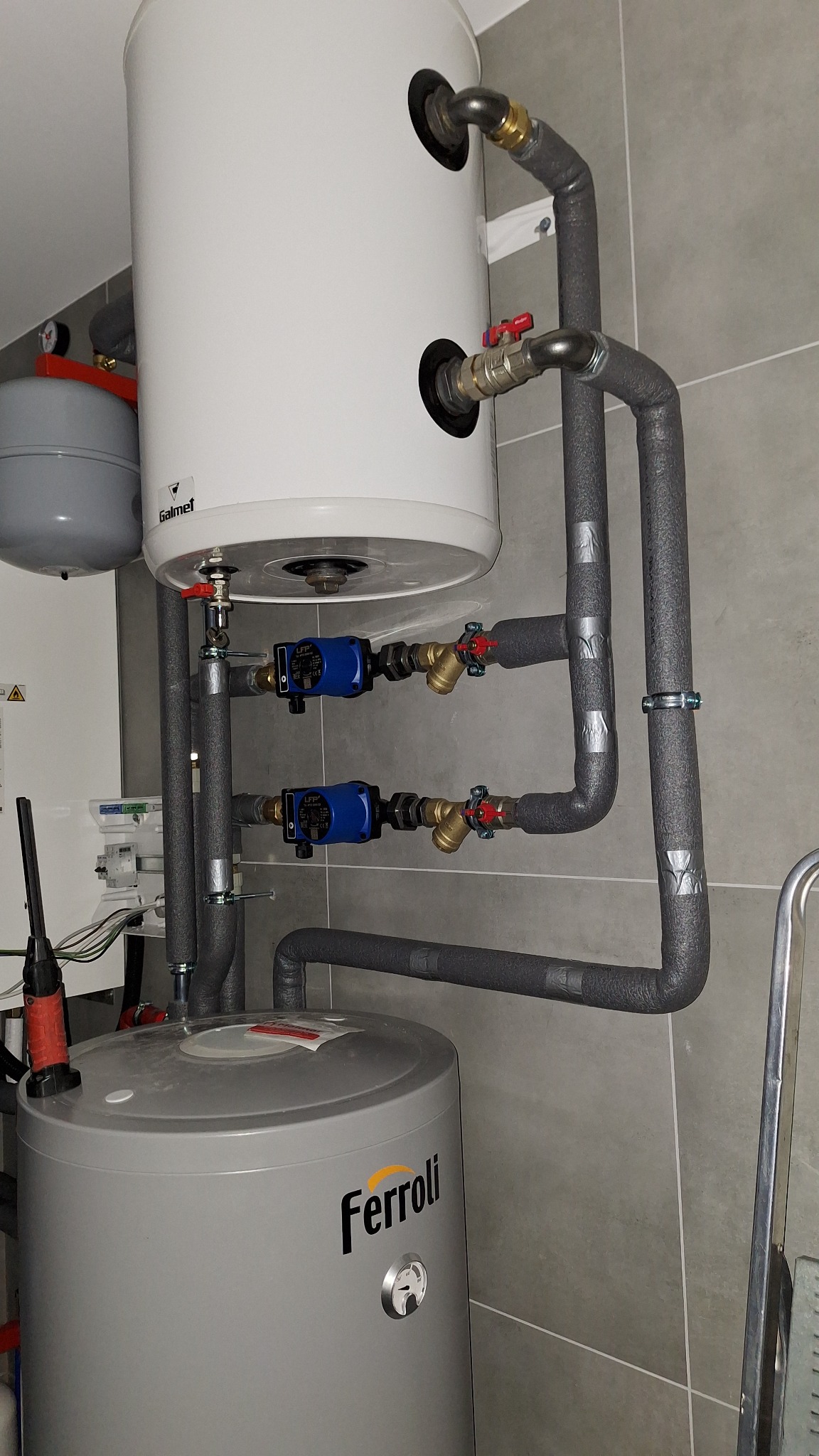 Instalacja dwóch zasobników ciepłej wody (Galmet i Ferroli) z pompami obiegowymi, zaworami i izolowanymi rurami w kotłowni, na tle szarych płytek.