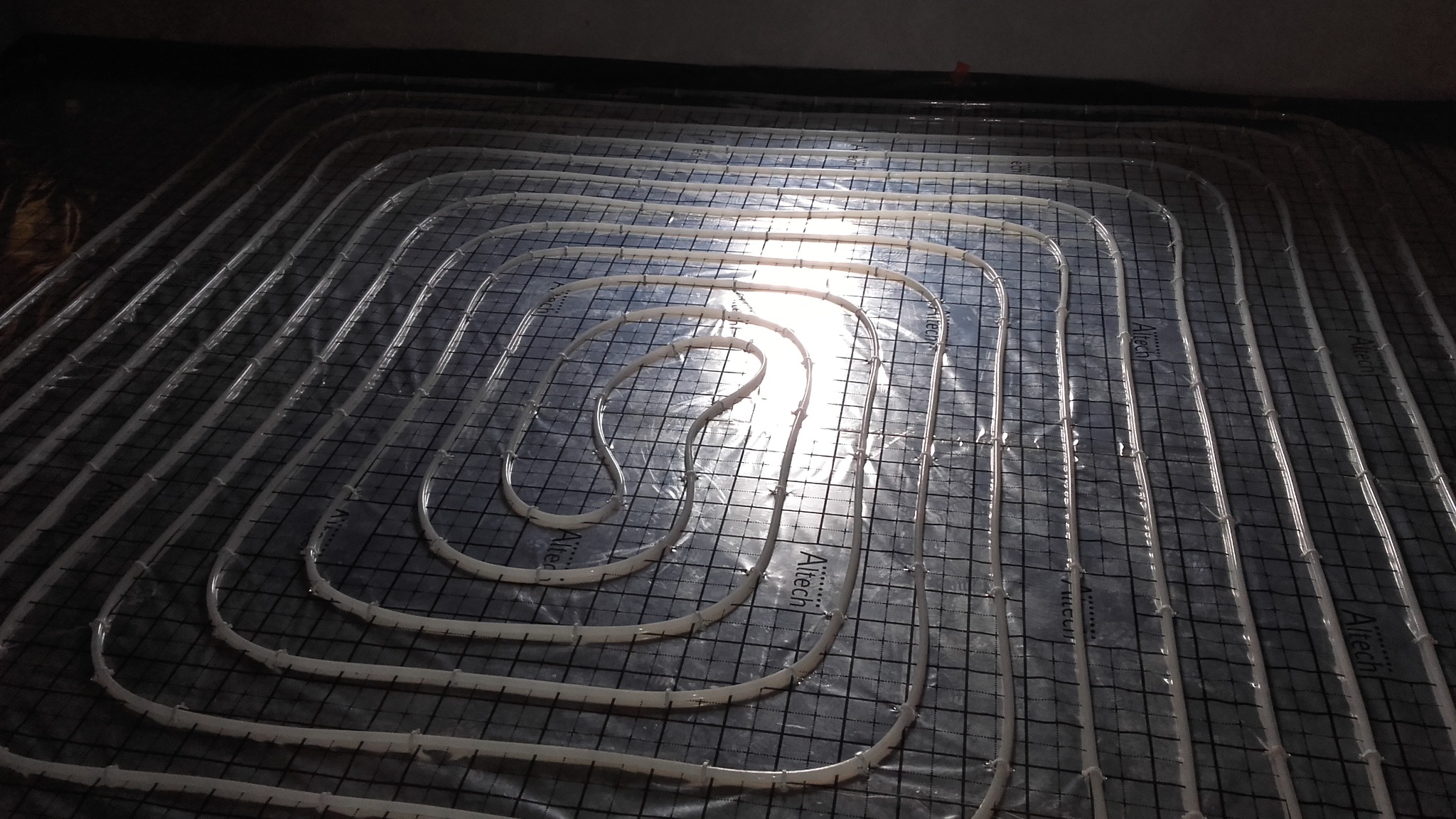 Ułożone rury ogrzewania podłogowego na czarnej folii z siatką, widok z góry, charakterystyczny wzór spiralny w centrum, instalacja Aitech.