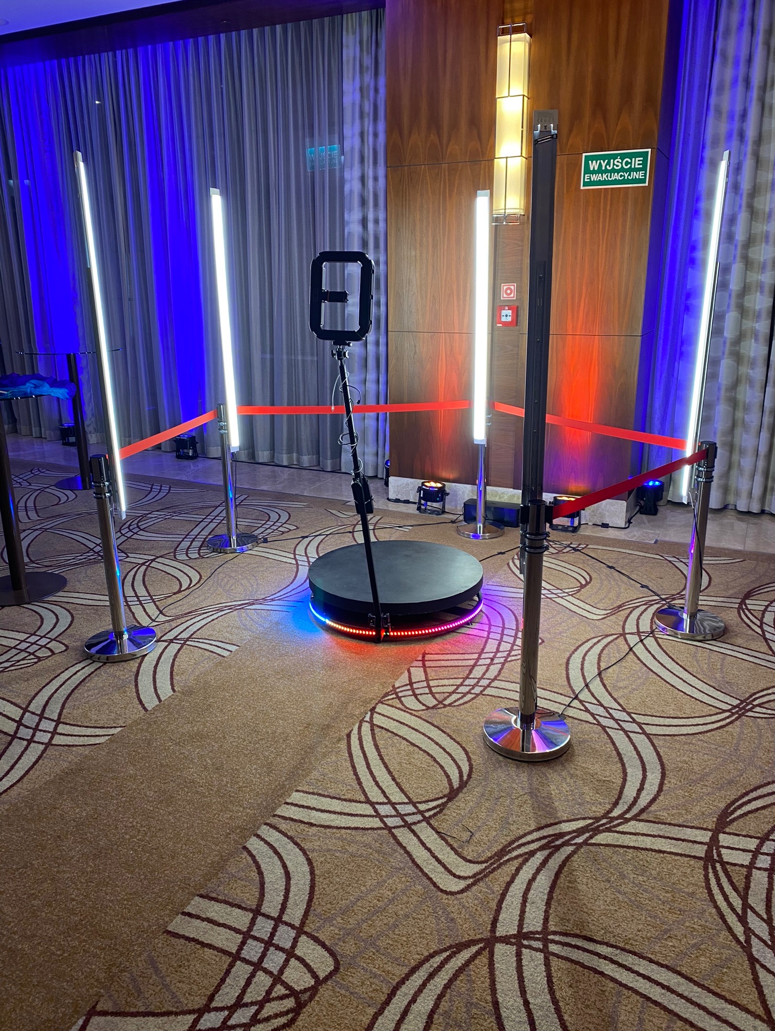 Rotacyjna platforma 360° z oświetleniem LED, otoczona słupkami z taśmą. Tło stanowią zasłony i drewniana ściana z lampami. Dekoracja imprezowa.