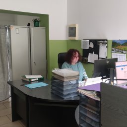 Biuro rachunkowe Ewa&Ewa - Biuro: kobieta siedzi przy biurku z komputerami i dokumentami. Widoczny kalendarz, szafa i zasłony. Stonowana kolorystyka ścian.