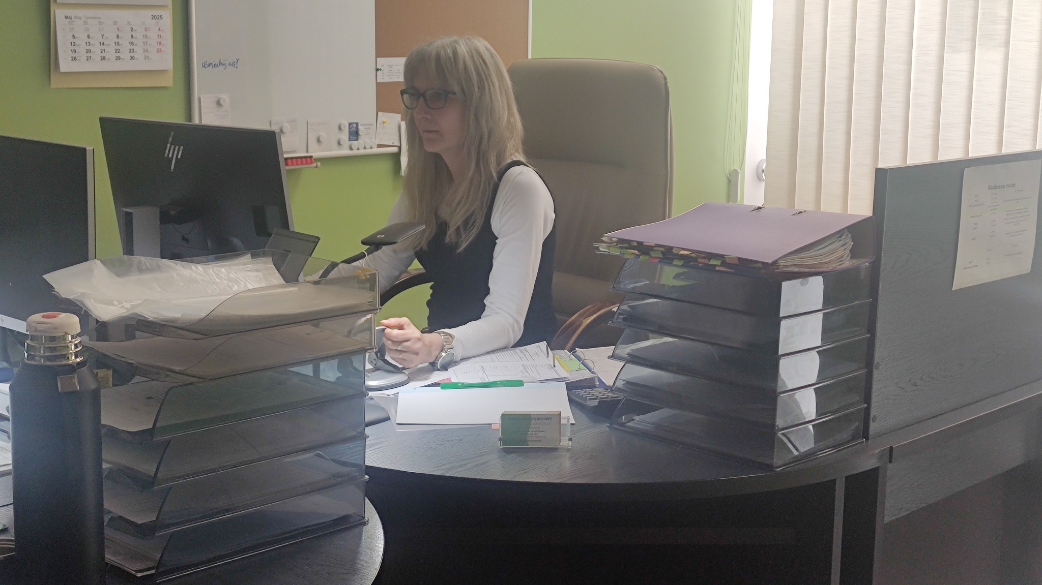 Kobieta w okularach siedzi przy biurku w biurze, na którym leżą dokumenty i segregatory. W tle kalendarz i tablica.