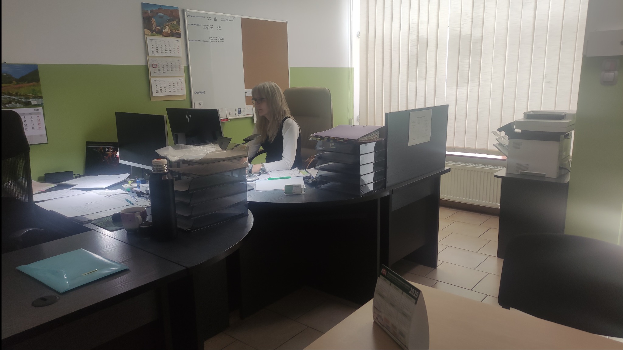 Kobieta przy biurku w biurze, otoczona stertami dokumentów, komputerem i kalendarzem. W tle okno z żaluzjami i tablica suchościeralna.