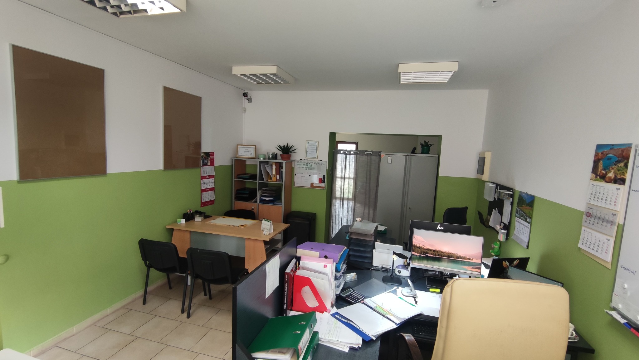 Wnętrze biura rachunkowego: biurka z komputerami, segregatory, kalendarze na ścianach, zielona lamperia, widok na stanowiska pracy.