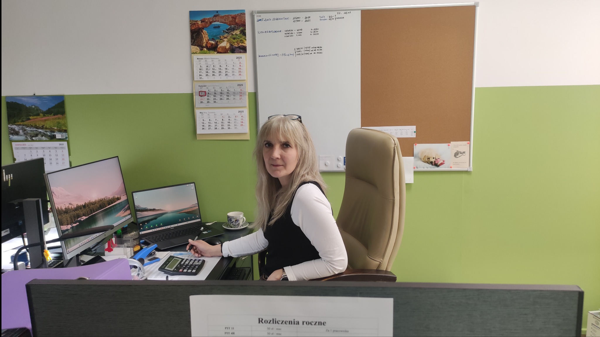 Kobieta przy biurku z kalkulatorem i długopisem, w tle monitory, kalendarze i tablica informacyjna. Rozliczenia roczne na pierwszym planie.