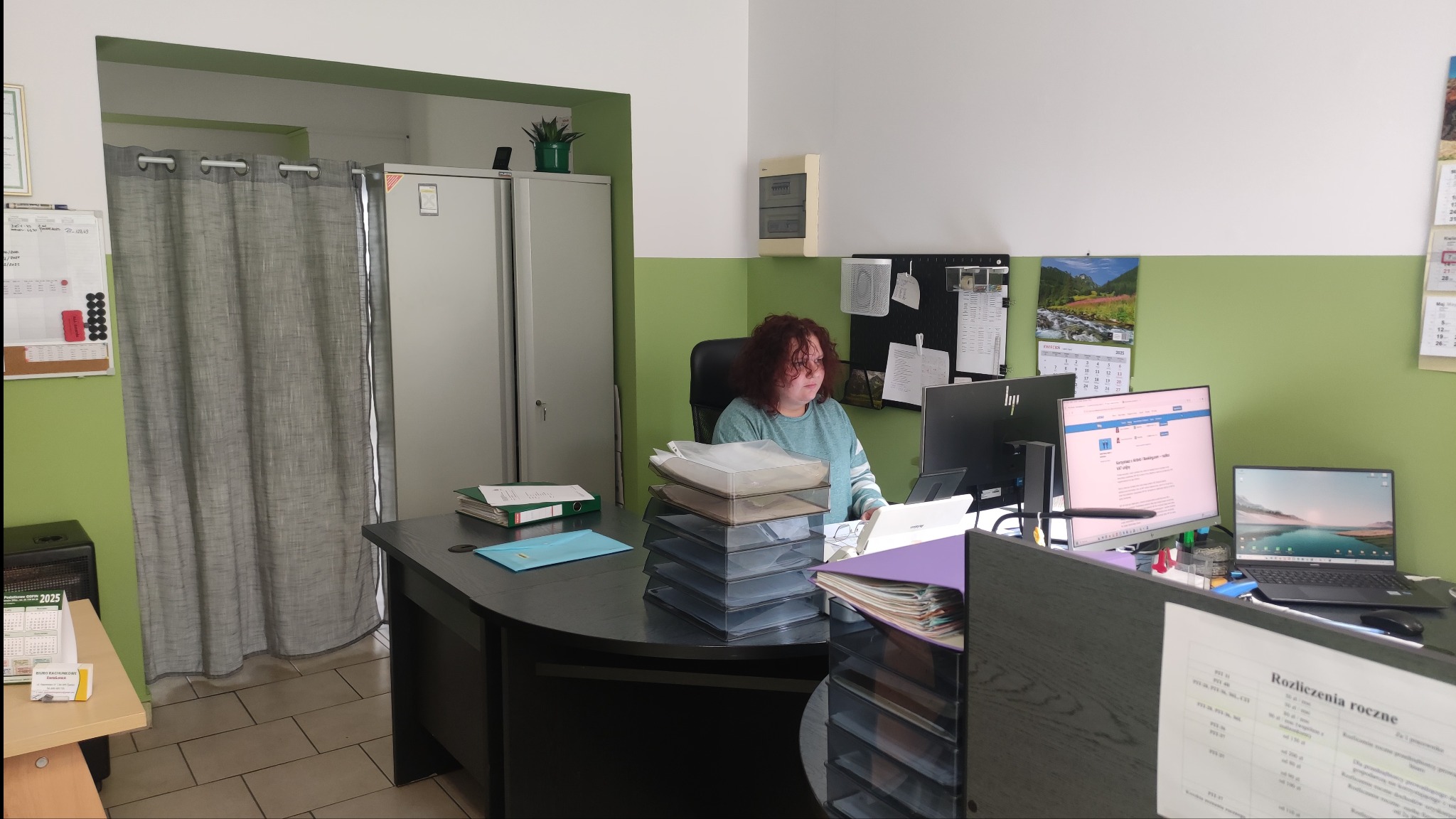 Biuro: kobieta siedzi przy biurku z komputerami i dokumentami. Widoczny kalendarz, szafa i zasłony. Stonowana kolorystyka ścian.