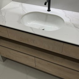 ANDRII KOLTSOV STREAMCO MEBLE - Nowoczesna szafka łazienkowa z drewnianymi frontami, białym blatem imitującym marmur i czarną armaturą. Nad nią podświetlane lustro.