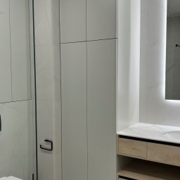 ANDRII KOLTSOV STREAMCO MEBLE - Jasna łazienka z zabudową meblową: szafa wnękowa z szarymi frontami i drewniana szafka pod umywalkę, białe płytki imitujące marmur, kabina prysznicowa.