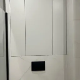 Białe szafki na wymiar w łazience nad toaletą, z czarnym przyciskiem spłuczki i marmurowymi płytkami na ścianie. Minimalistyczny design.