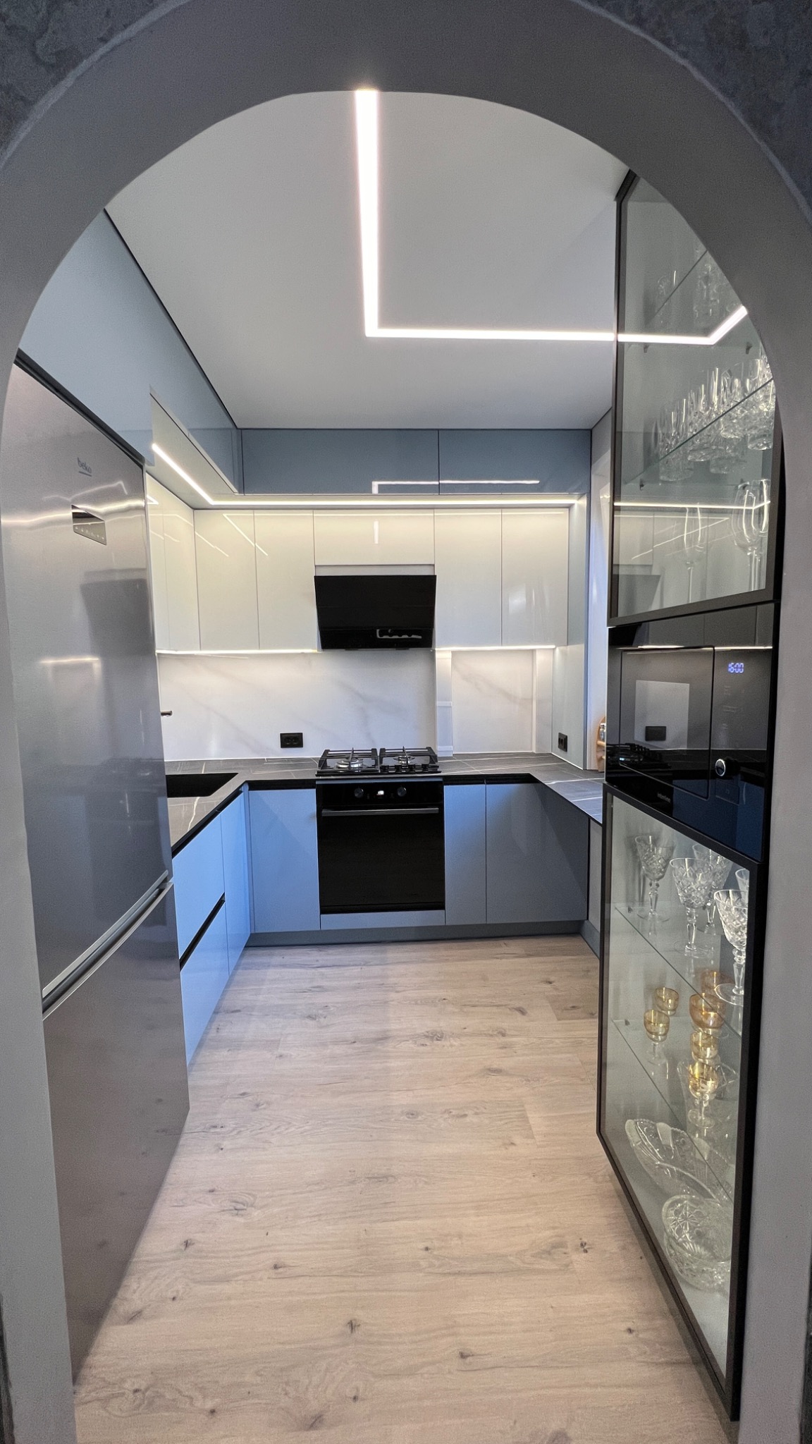 Nowoczesna, jasna kuchnia w odcieniach szarości i bieli, z subtelnym oświetleniem LED i przeszkloną witryną na zastawę, widziana przez kamienny łuk.