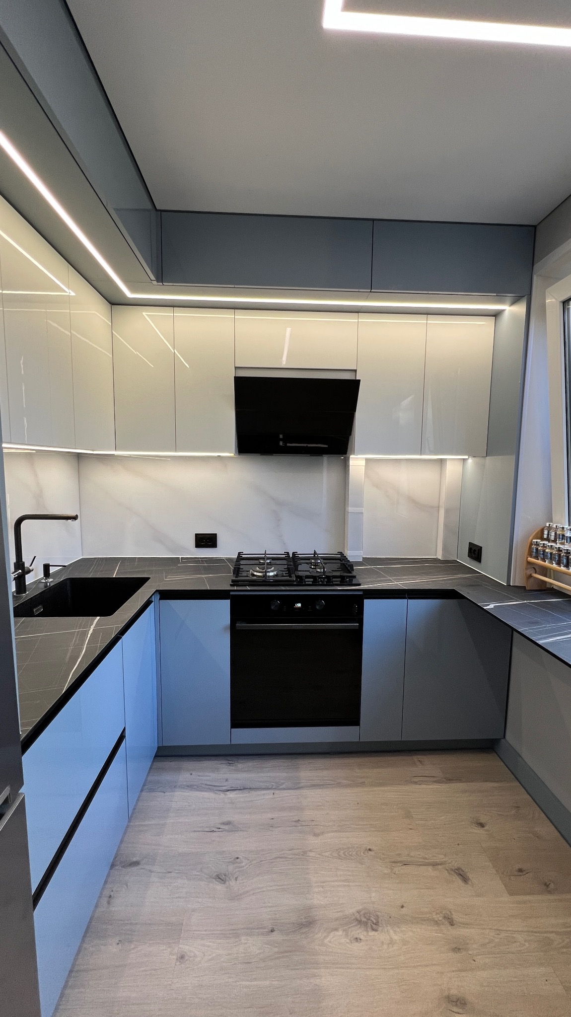 Nowoczesna kuchnia z szarymi szafkami, czarnym blatem imitującym marmur i oświetleniem LED. Widoczna płyta gazowa i okap. Jasna podłoga.
