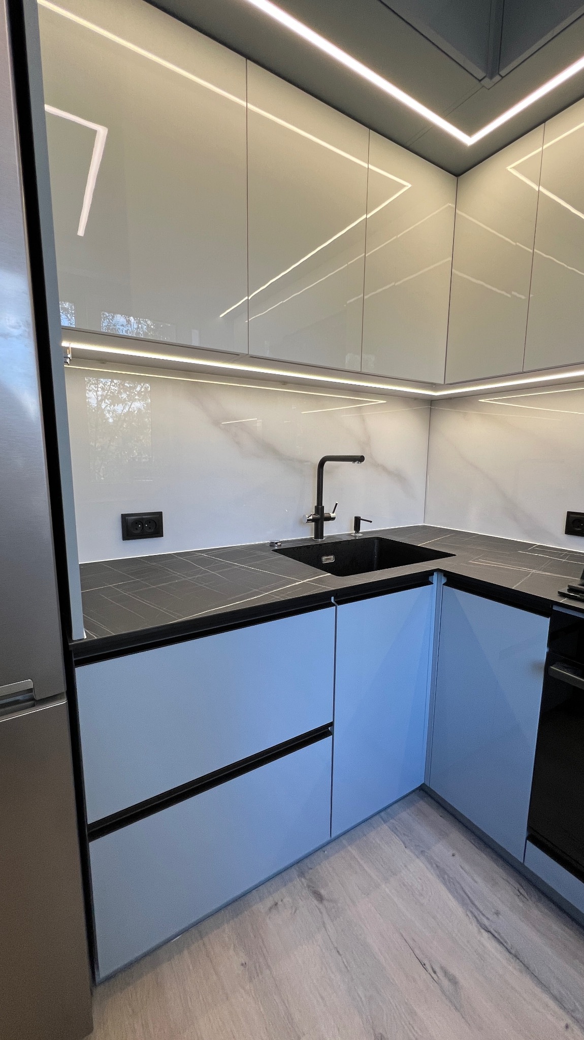 Nowoczesna kuchnia z szarymi szafkami, czarnym blatem i eleganckim oświetleniem LED. Widoczny zlew i bateria w czarnym kolorze.