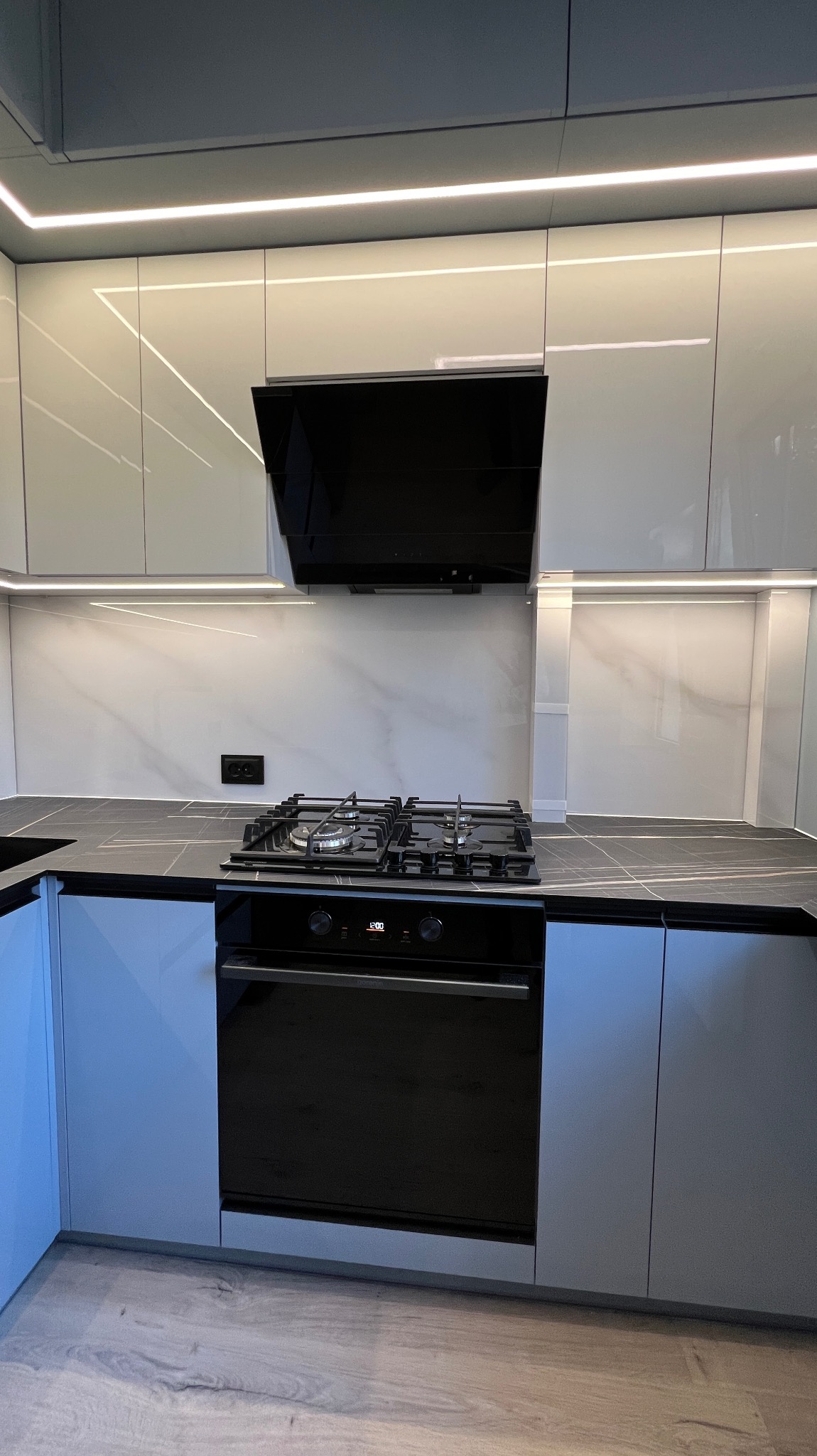 Nowoczesna kuchnia na wymiar z szarymi szafkami, czarnym blatem i sprzętem AGD. Oświetlenie LED podkreśla minimalistyczny design i marmurowy wzór ściany.