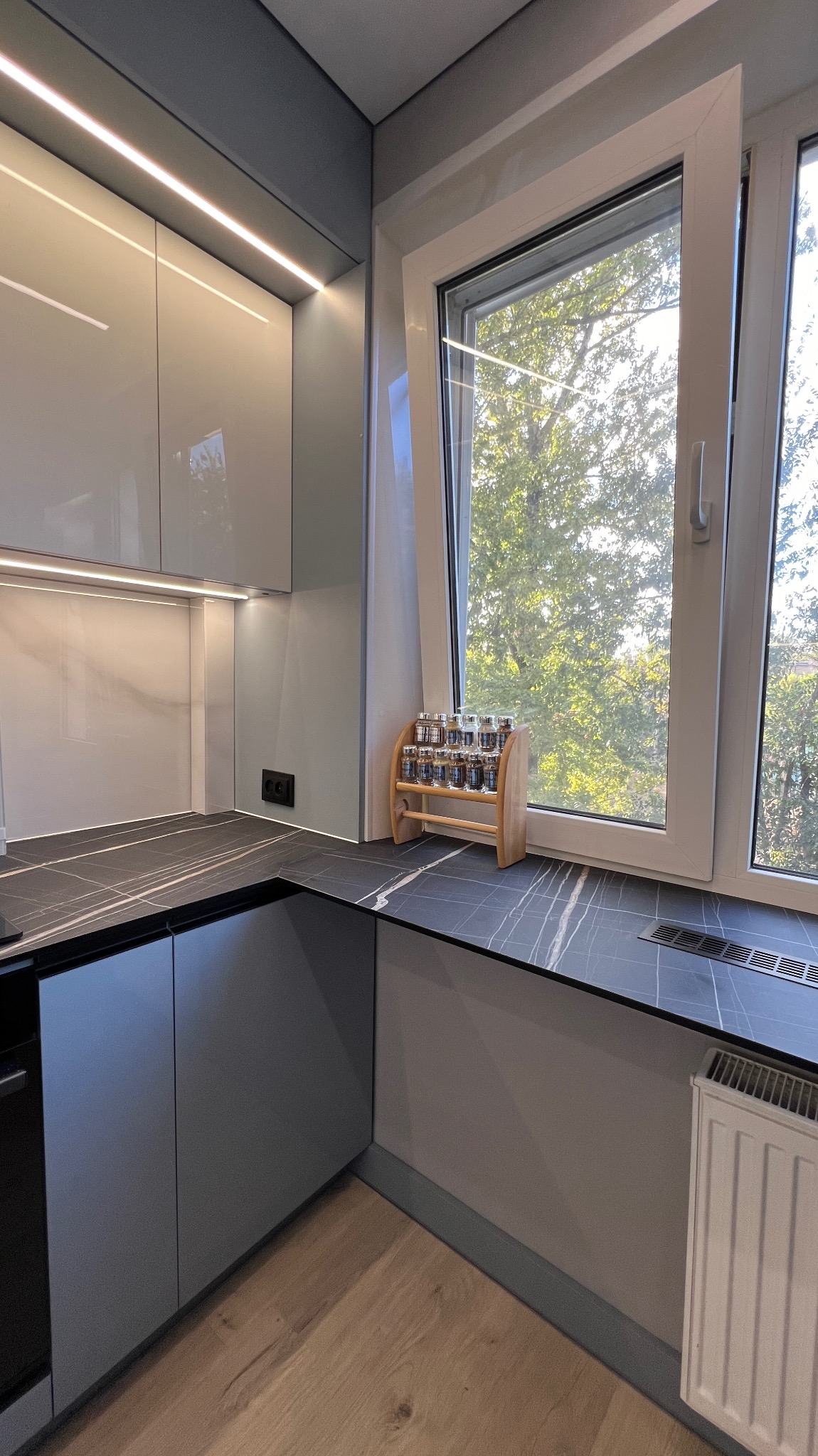 Nowoczesna kuchnia: szare szafki z połyskiem, blat imitujący marmur, okno z widokiem na zieleń. Drewniana półka na przyprawy dodaje ciepła wnętrzu.