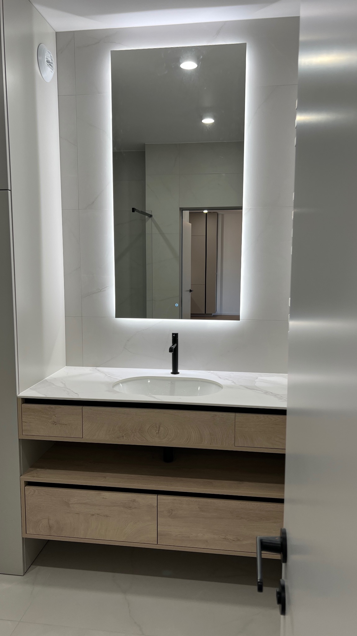 Nowoczesna łazienka z podświetlanym lustrem i szafką z drewnianymi frontami. Widoczna armatura w kolorze czarnym i marmurowy blat. Minimalistyczny design.