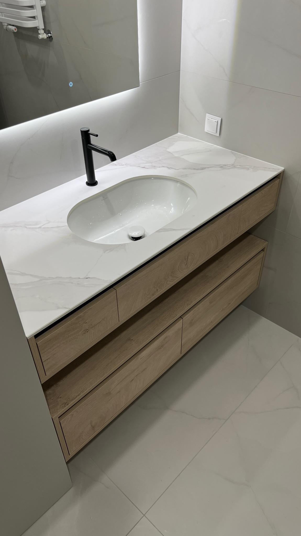 Nowoczesna łazienka: biały blat z umywalką, drewniane szafki z szufladami, czarna bateria, lustro z podświetleniem LED i marmurowe płytki podłogowe.