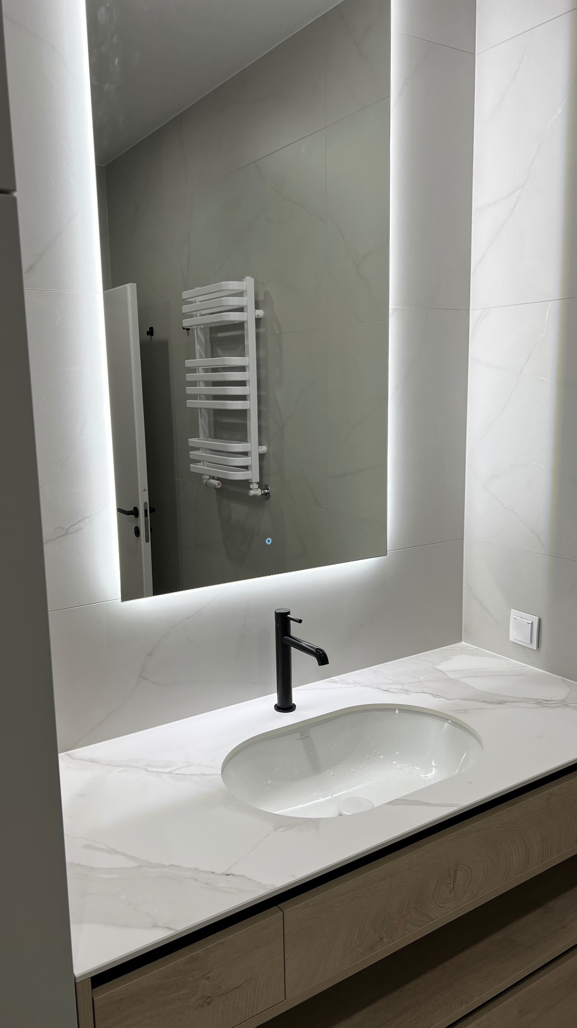 Nowoczesna łazienka z umywalką wpuszczaną w marmurowy blat, podświetlane lustro i grzejnik drabinkowy. Minimalistyczny design, jasne kolory.