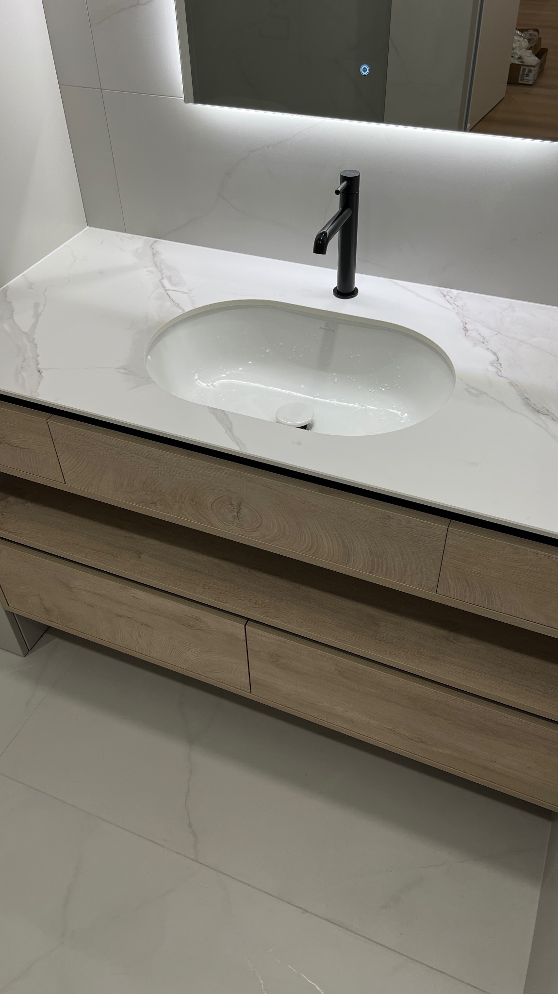 Nowoczesna szafka łazienkowa z drewnianymi frontami, białym blatem imitującym marmur i czarną armaturą. Nad nią podświetlane lustro.