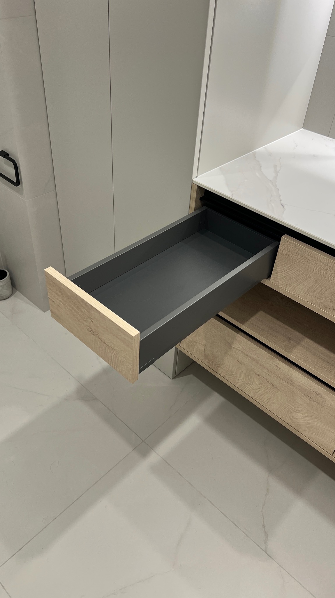 Wysuwana szuflada z drewnianym frontem w zabudowie kuchennej z białym blatem i szarymi frontami, widok z boku, na tle jasnych płytek podłogowych.