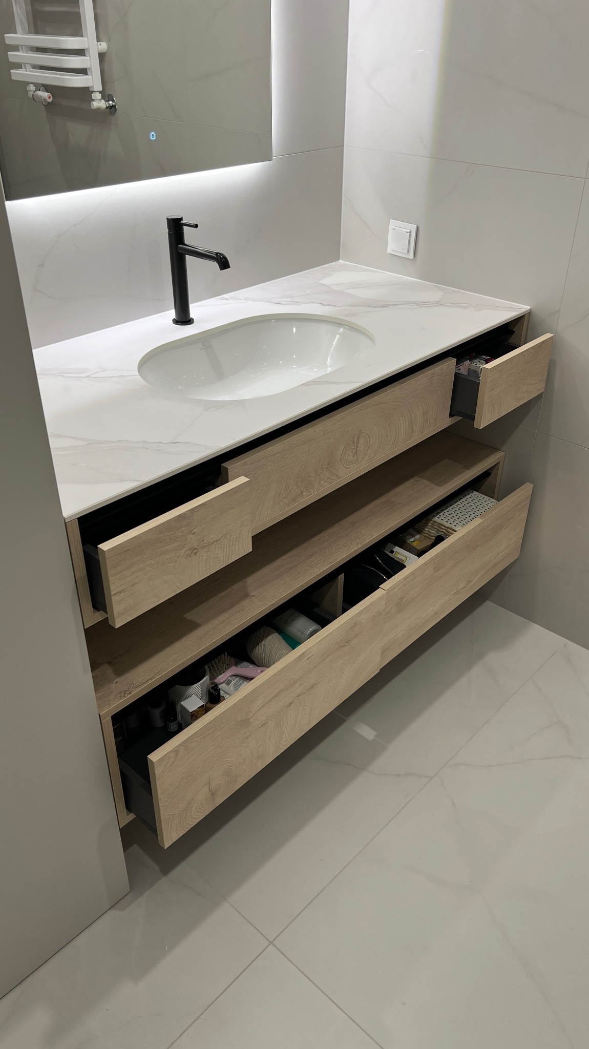 Nowoczesna szafka łazienkowa z blatem imitującym marmur, czarną armaturą i otwartymi szufladami z widoczną zawartością. Minimalistyczny design, jasne płytki.